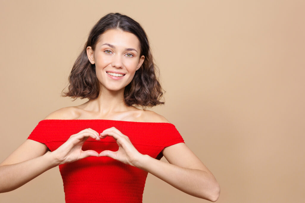 woman on a beige background smiles and makes a heart symbol with her hands. a happy person rejoices